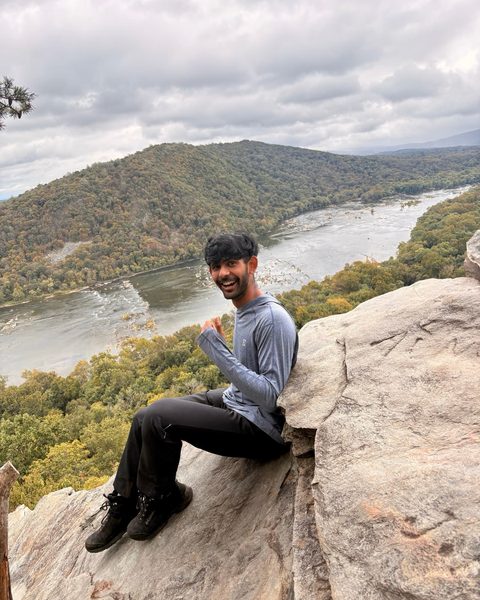 Harsh Patel seated on a rocky overlook above a river and forested hills